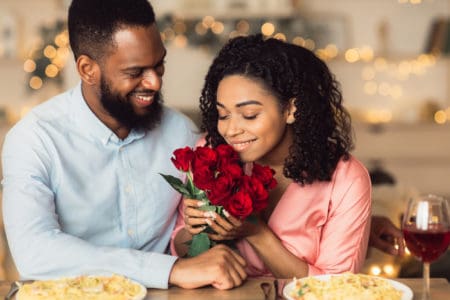 Greeting With Holidays. Happy beautiful black girlfriend receiving bouquet of red roses from her smiling boyfriend, smelling bunch of flowers during romantic date in luxury restaurant
